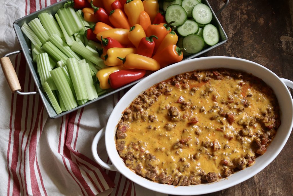 Cheesy baked taco dip in a oval white baking dish, a red and white striped hand towel and a tray of veggies for dipping.