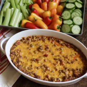 Cheesy baked taco dip in an oval white baking dish. Served with a side of raw vegetables. There's a red and white striped hand towel on the side.