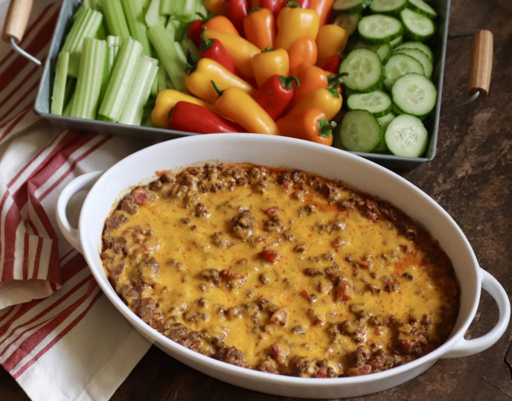 Cheesy baked taco dip in an oval white baking dish. Served with a side of raw vegetables. There's a red and white striped hand towel on the side.