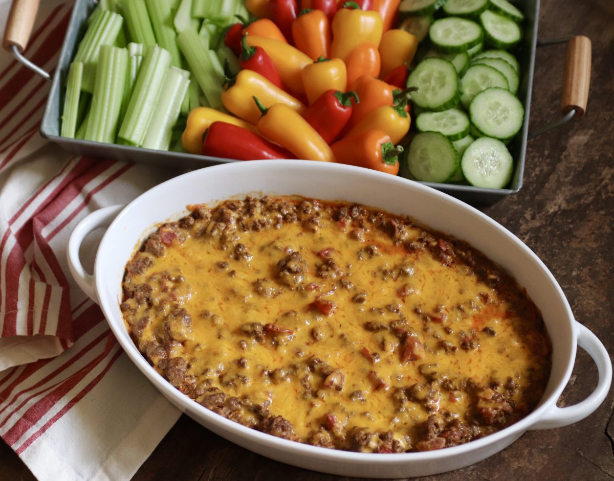 Cheesy baked taco dip in an oval white baking dish.  Served with a side of raw vegetables. There's a red and white striped hand towel on the side.  