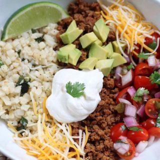 A burrito bowl with cauliflower lime rice, taco meat, shredded cheese, avocado, salsa and a lime wedge.