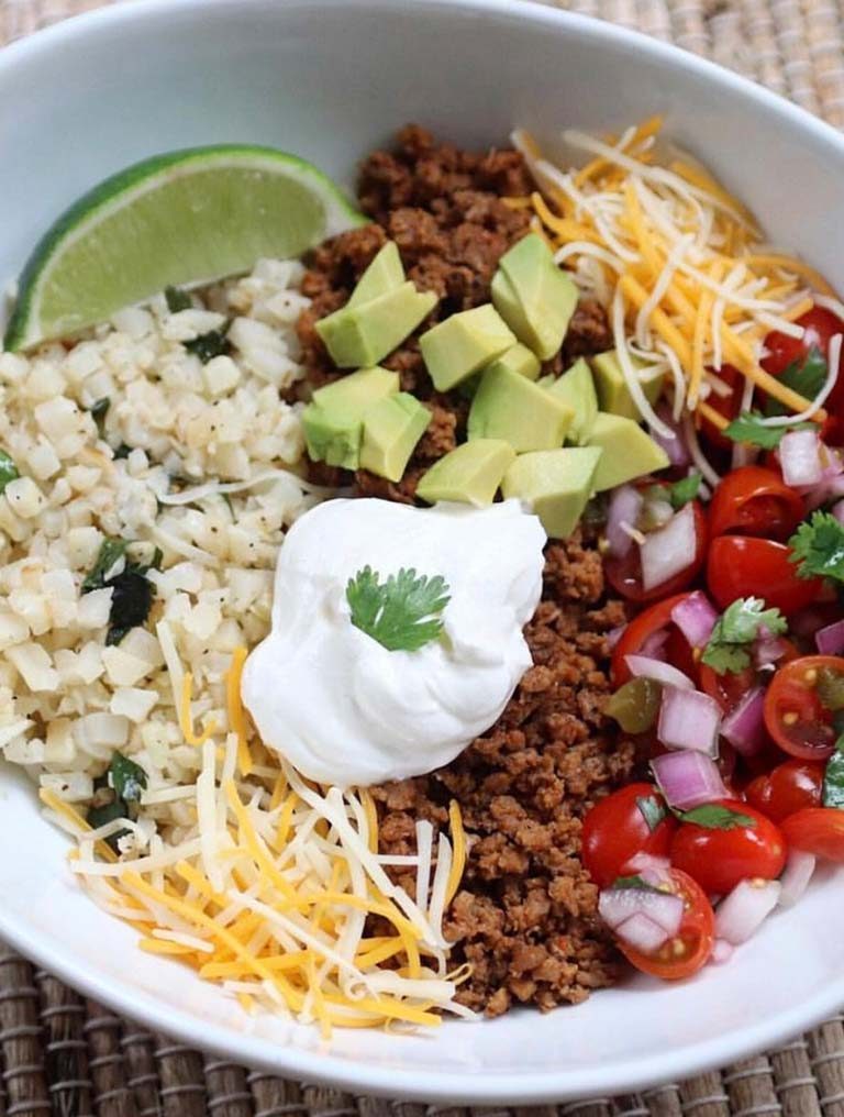 A burrito bowl with cauliflower lime rice, taco meat, shredded cheese, avocado, salsa and a lime wedge.