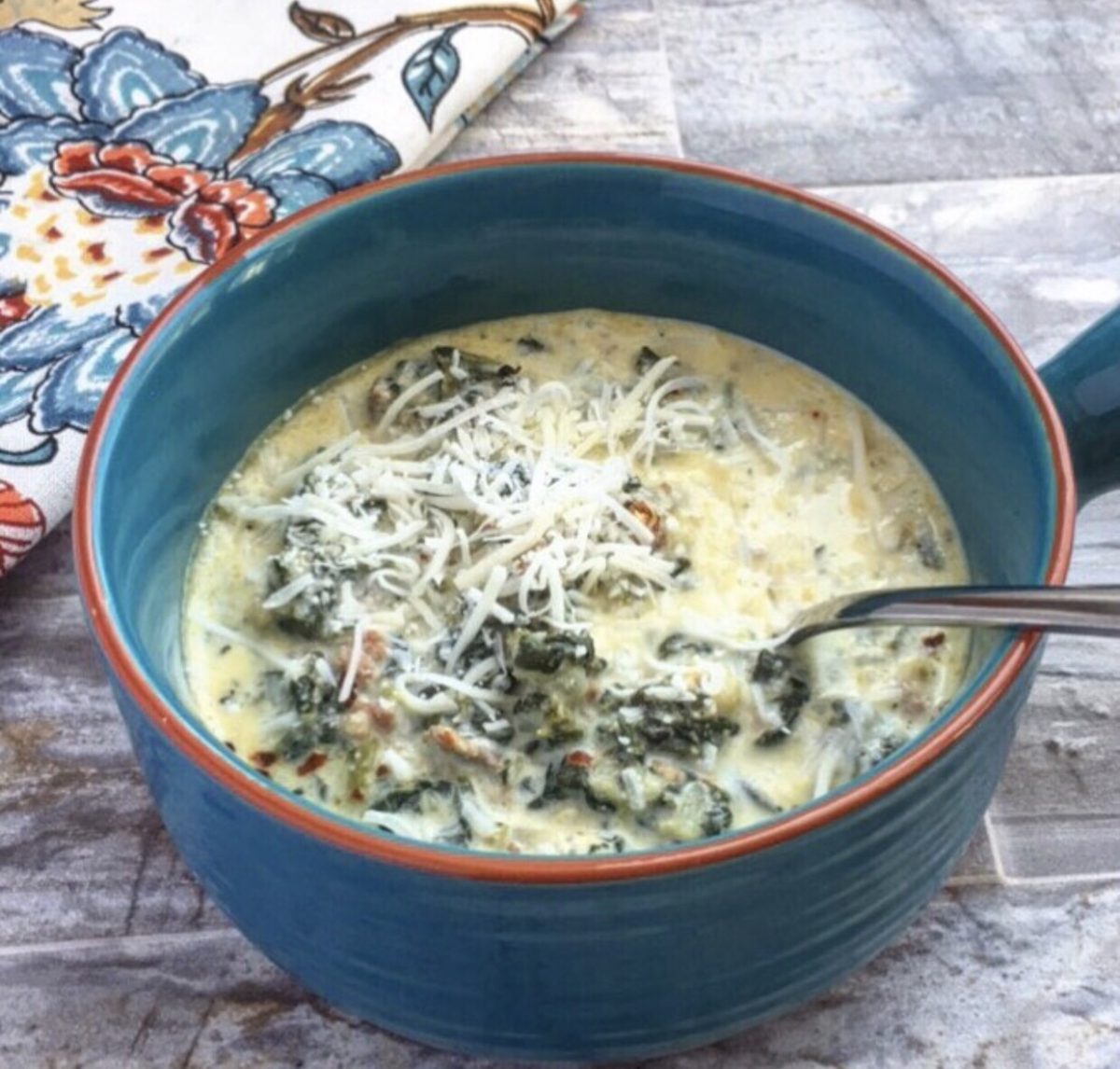 a blue stoneware bowl with creamy zuppa Toscana soup in it. a floral blue and orange napkin beside it.