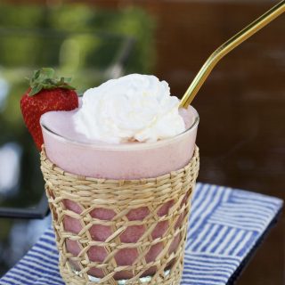 A strawberry banana smoothie in a glass with a rattan sleeve. It has a gold straw and is topped with whipped cream and garnished with a strawberry.