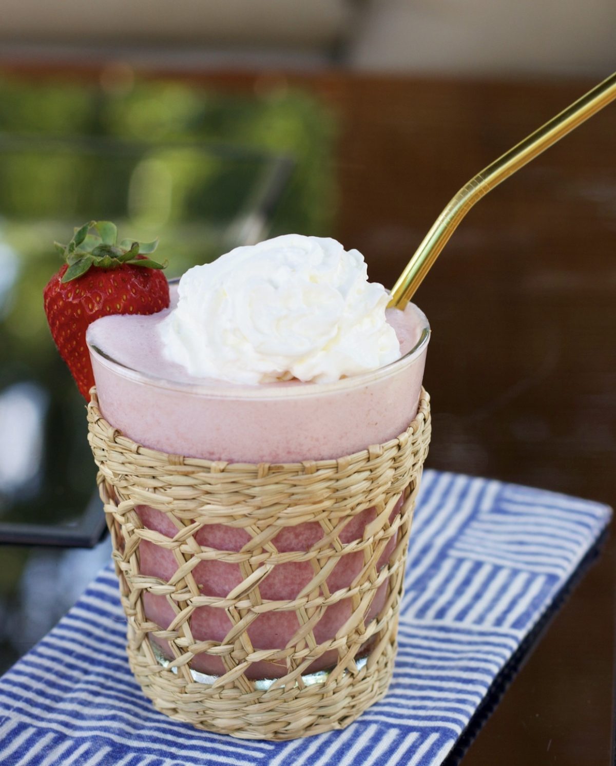 A strawberry banana smoothie in a glass with a rattan sleeve.  It has a gold straw and is topped with whipped cream and garnished with a strawberry.