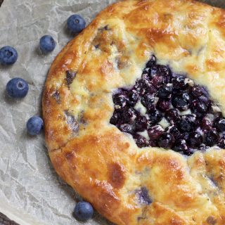 Golden Brown Blueberry galette on a brown piece of parchment paper. There's a few blueberries on the parchment paper.