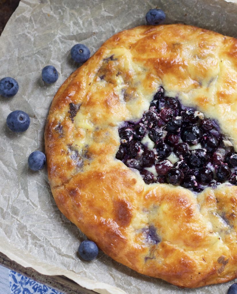 Golden Brown Blueberry galette on a brown piece of parchment paper. There's a few blueberries on the parchment paper.