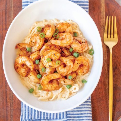 A white bowl of Alfredo pasta topped with spicy cajun shrimp and garnished with green onions. It's sitting on a blue and white geometric napkin, on top of a round dark wood charger plate. There's a gold fork to eat it with.