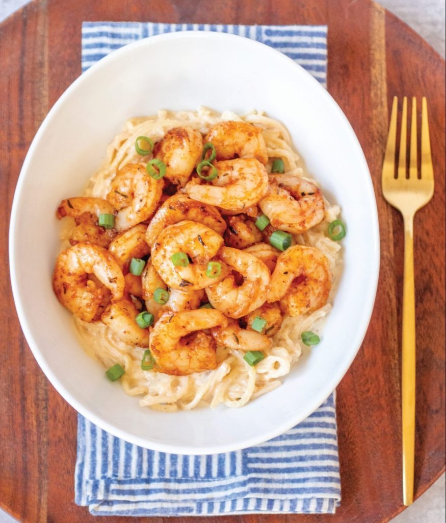 A white bowl of Alfredo pasta topped with spicy cajun shrimp and garnished with green onions. It's sitting on a blue and white geometric napkin, on top of a round dark wood charger plate. There's a gold fork to eat it with.