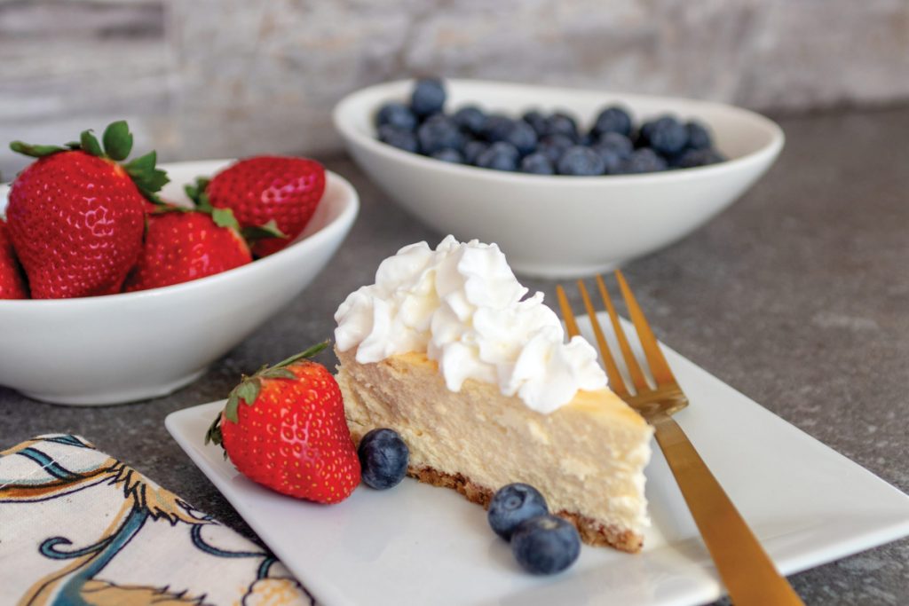 A slice of cheesecake on a square white plate with a gold fork. Strawberries and blueberries for garnish.