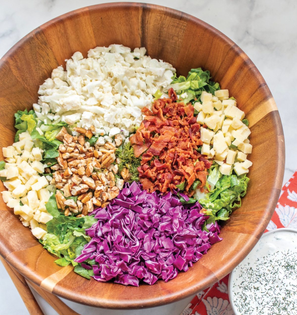 A wood bowl with salad ingredients, red cabbage, romaine lettuce, cauliflower, pecans, bacon and cheese. Beside the bowl are wood salad tongs and a coral napkin.