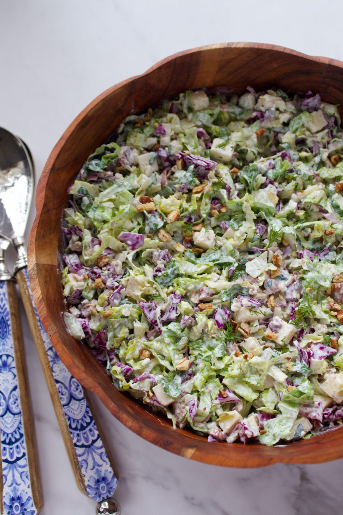 A large wooden salad bowl with a colorful dill pickle chopped salad in it. The serving tongs are blue and white with silver and wood accents.