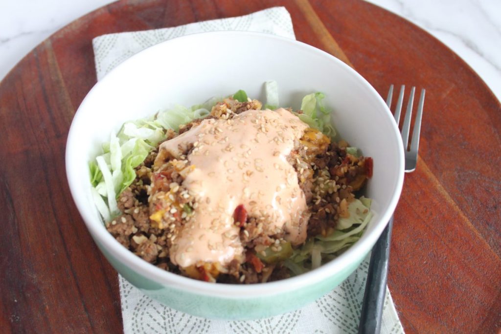 A green and white bowl of keto Big Mac skillet on top of iceberg lettuce. The bowl is sitting on a cloth napkin on a dark wood charger plate.