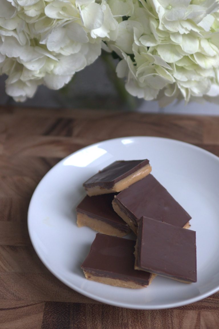 A white plate with peanut butter chocolate bars on it. The plate is sitting on a wood block. White hydrangea flowers are in the backdrop of the photo.