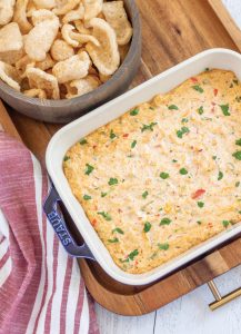 Creamy spicy keto corn dip in a blue ceramic stub baking dish. Served with a bowl of pork rinds. The dip and bowl are sitting on a wood tray. There's a red and white striped hand towel on the left side.