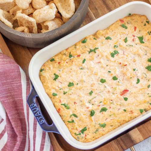 Creamy spicy keto corn dip in a blue ceramic stub baking dish. Served with a bowl of pork rinds. The dip and bowl are sitting on a wood tray. There's a red and white striped hand towel on the left side.