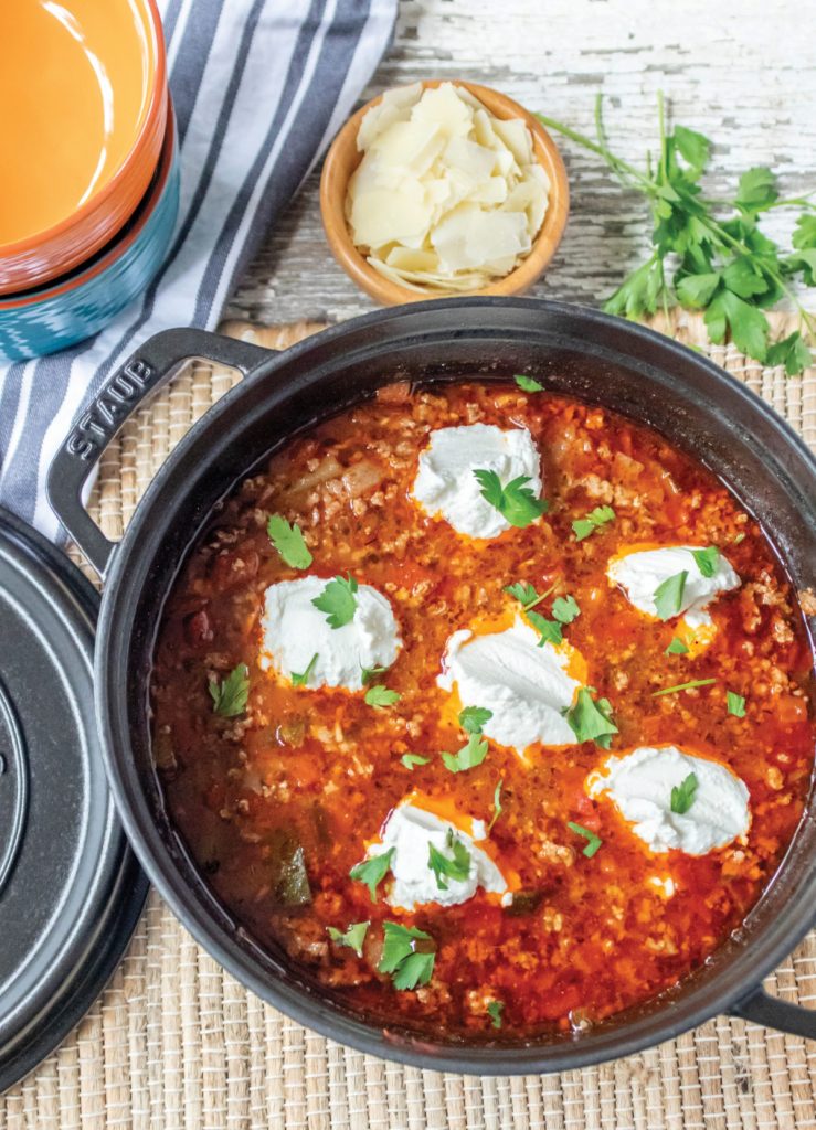 Lasagna soup in a black Staub pot. Shaved parmesan cheese in a round wood bowl. Parsley for garnish. Bowls for serving and a striped hand towel.