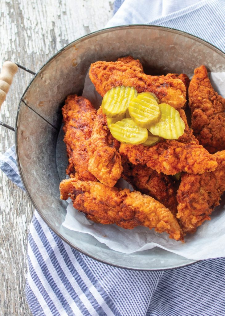 Nashville hot chicken tenders in a round metal serving container. It's sitting on a blue and white stripped hand towel. The chicken tenders have dill pickle slices on top of them.