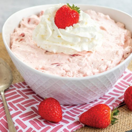 Strawberry Jello Fluff Salad in a white bowl, topped with whipped cream. The bowl is sitting on a graphic red and white hand towel. There are two strawberries by the bowl.