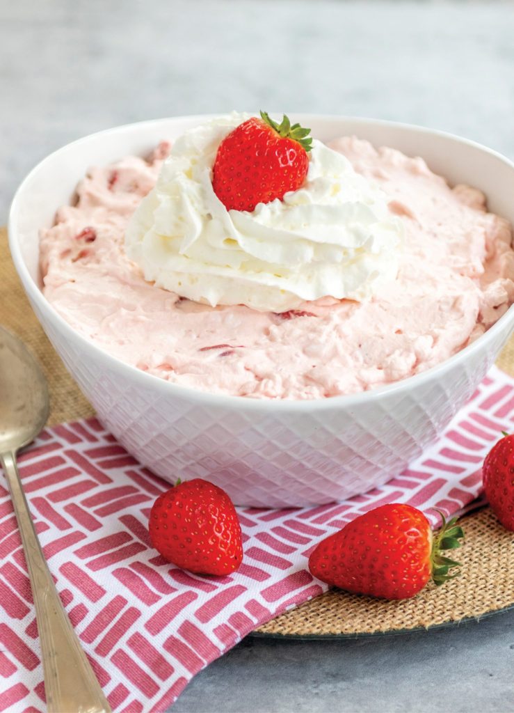 Strawberry Jello Fluff Salad in a white bowl, topped with whipped cream. The bowl is sitting on a graphic red and white hand towel. There are two strawberries by the bowl.