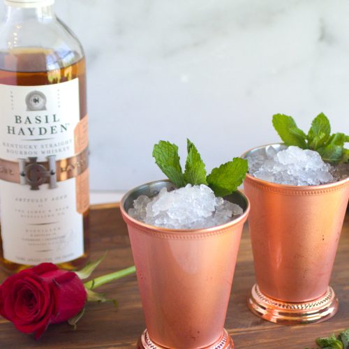 two mint juleps in copper julep cups. A red rose. A bottle of bourbon. the mint juleps are garnished with mint.