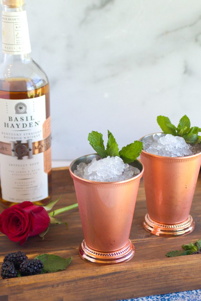 two mint juleps in copper julep cups. A red rose. A bottle of bourbon. the mint juleps are garnished with mint.