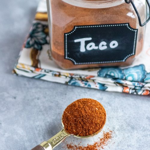 Taco seasoning in a labeled jar with a lid. The jar is sitting on a floral napkin. A tablespoon with taco season mix in it.