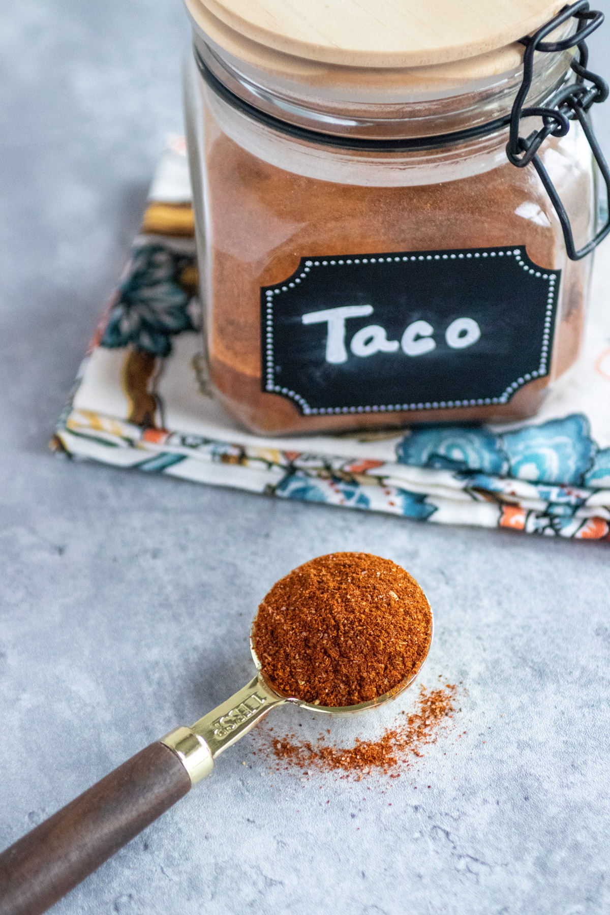 Taco seasoning in a labeled jar with a lid.  The jar is sitting on a floral napkin. A tablespoon with taco season mix in it.