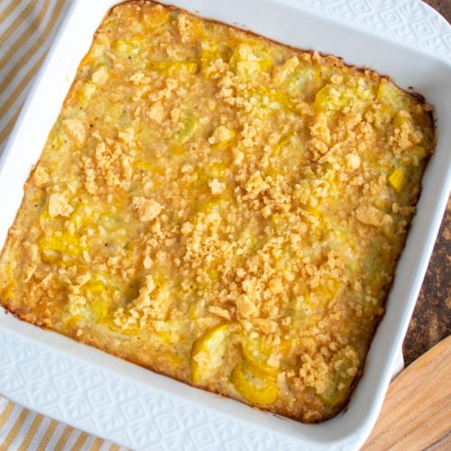 A square white casserole dish with squash casserole. The dish is sitting on a yellow and white striped napkin.