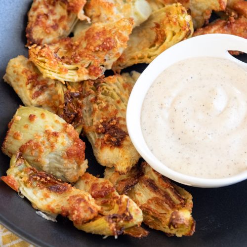 air fried artichoke hearts in a shallow black bowl. Garlic aoili for dipping in a small white bowl. a graphic yellow and white napkin underneath the bowl.