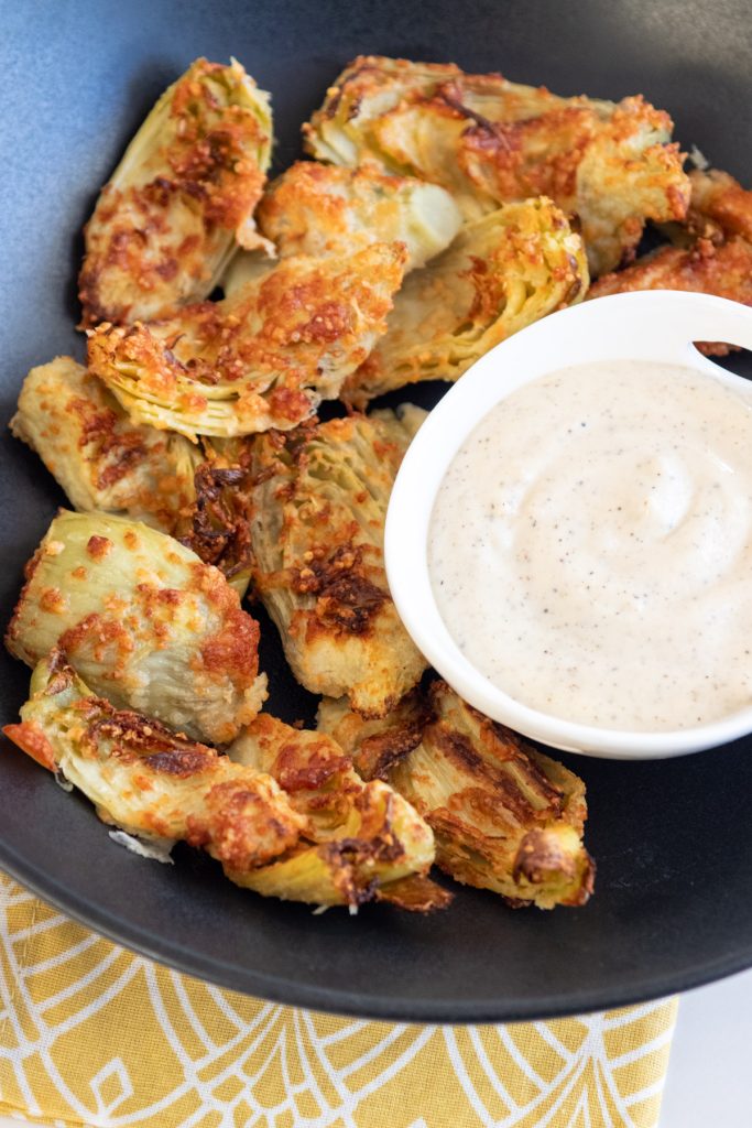 air fried artichoke hearts in a shallow black bowl. Garlic aoili for dipping in a small white bowl. a graphic yellow and white napkin underneath the bowl.