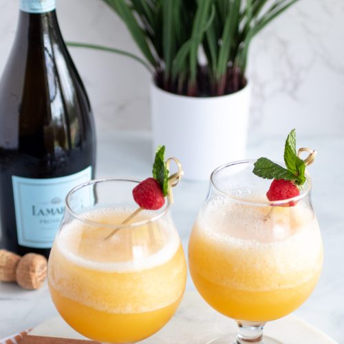 two frozen peach Bellinis. a bottle of Prosecco. a green plant behind the drinks and Prosecco.