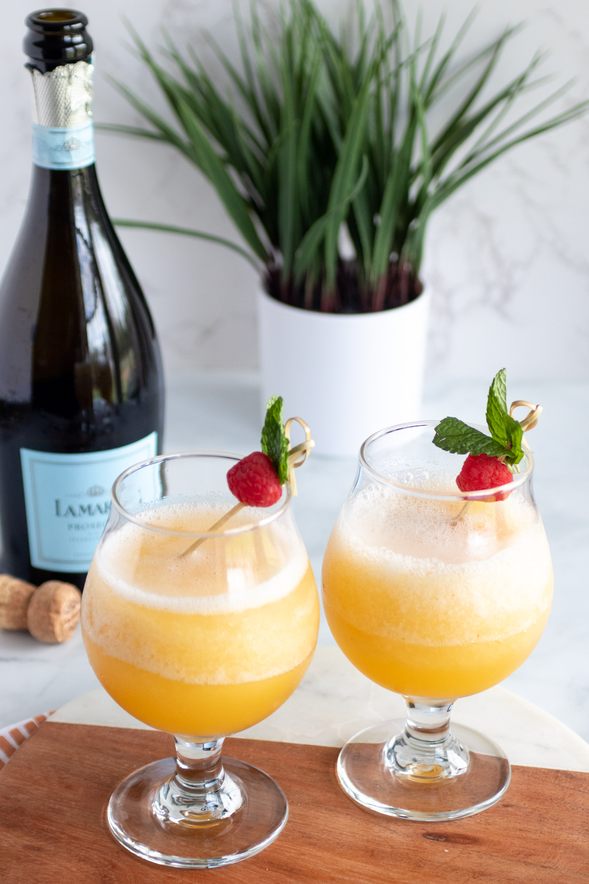 two frozen peach Bellinis. a bottle of Prosecco. a green plant behind the drinks and Prosecco.