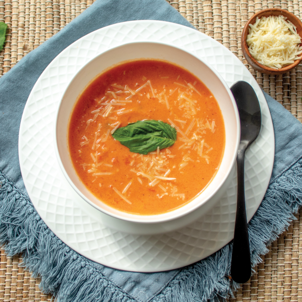 a white bowl with tomato basil soup. The soup is garnished with one basil leaf. The bowl is sitting on a white plate on a blue napkin. Black spoon to the right of plate. A small wood bowl of parmesan cheese for garnish.