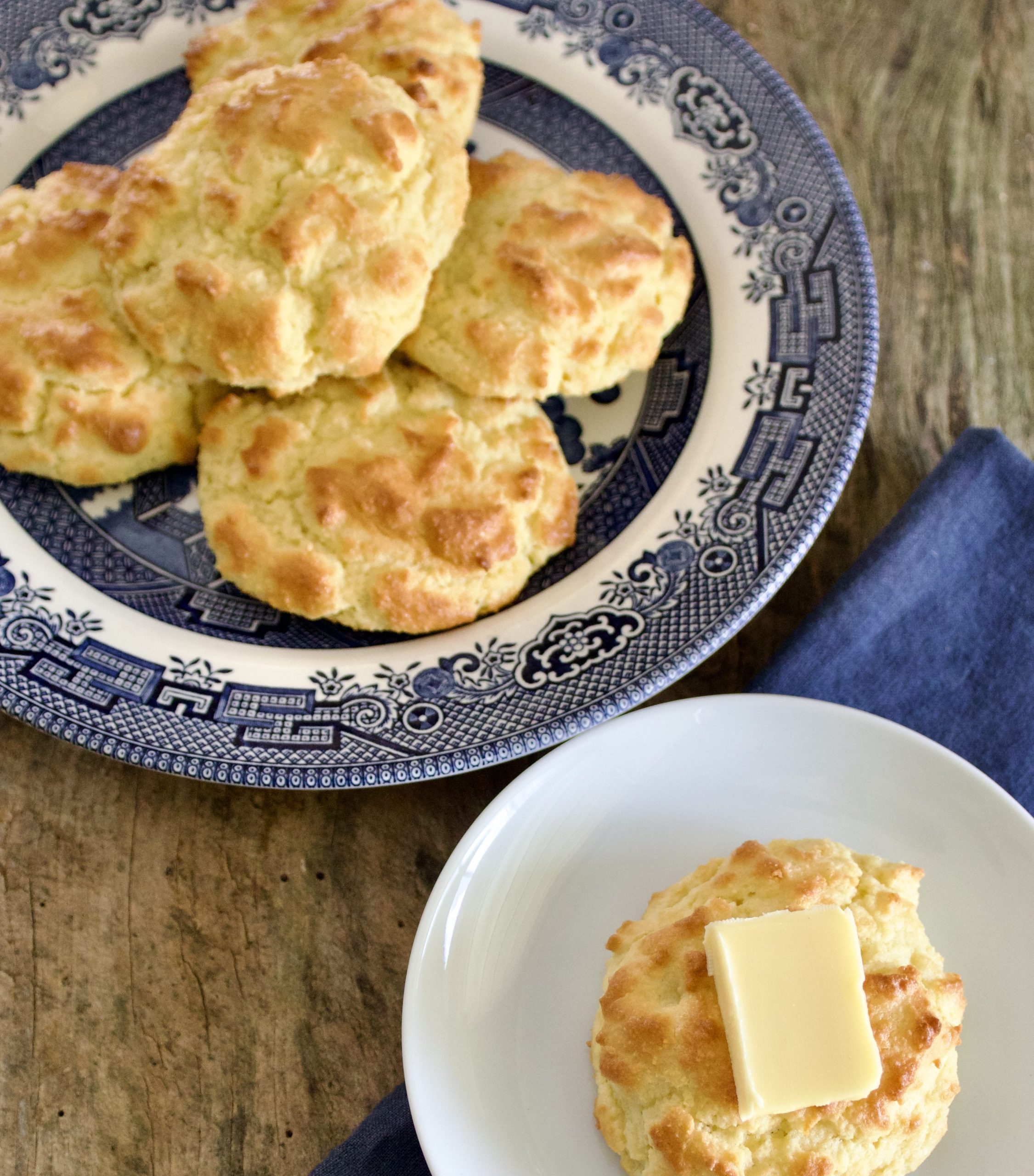 drop biscuits on a blue and white plate. one drop biscuit topped with a pat of butter on a small white plate. the plate is sitting on a blue napkin.