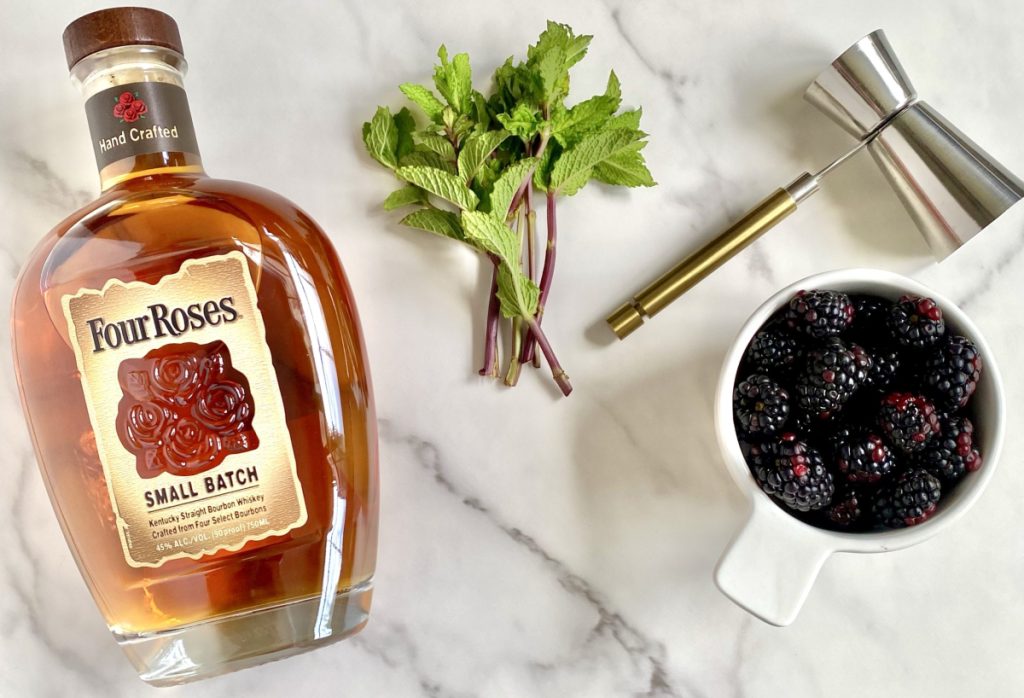 A bottle of four roses bourbon, mint leaves, a cup of blackberries and a  bar shot tool.