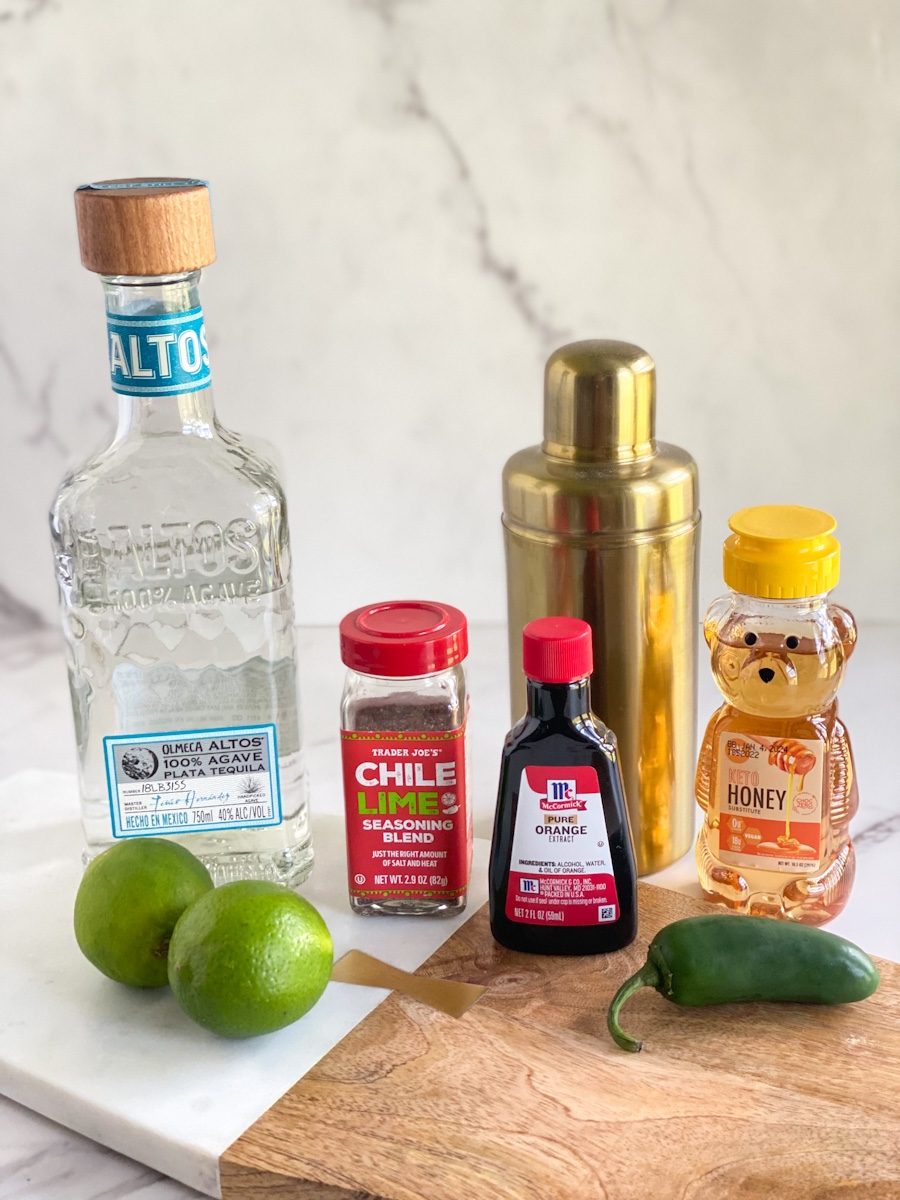 ingredients, tequila, jalapenos, limes, chili lime seasoning, keto honey, orange extract, and a gold cocktail shaker.