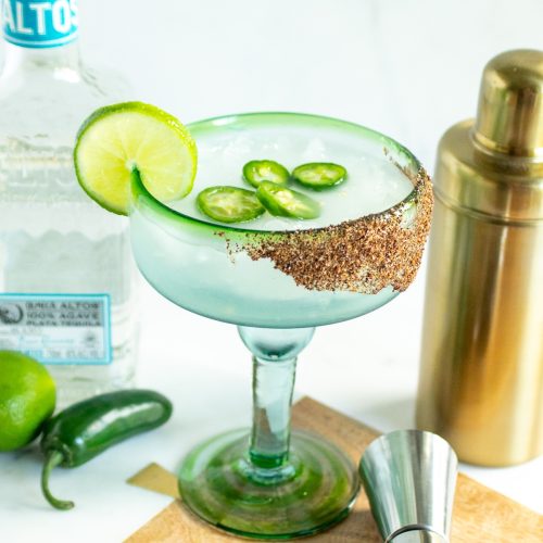 A margarita in a margarita-style glass garnished with jalapenos and lime. A bottle of tequila to the left. A gold cocktail shaker is to the right.