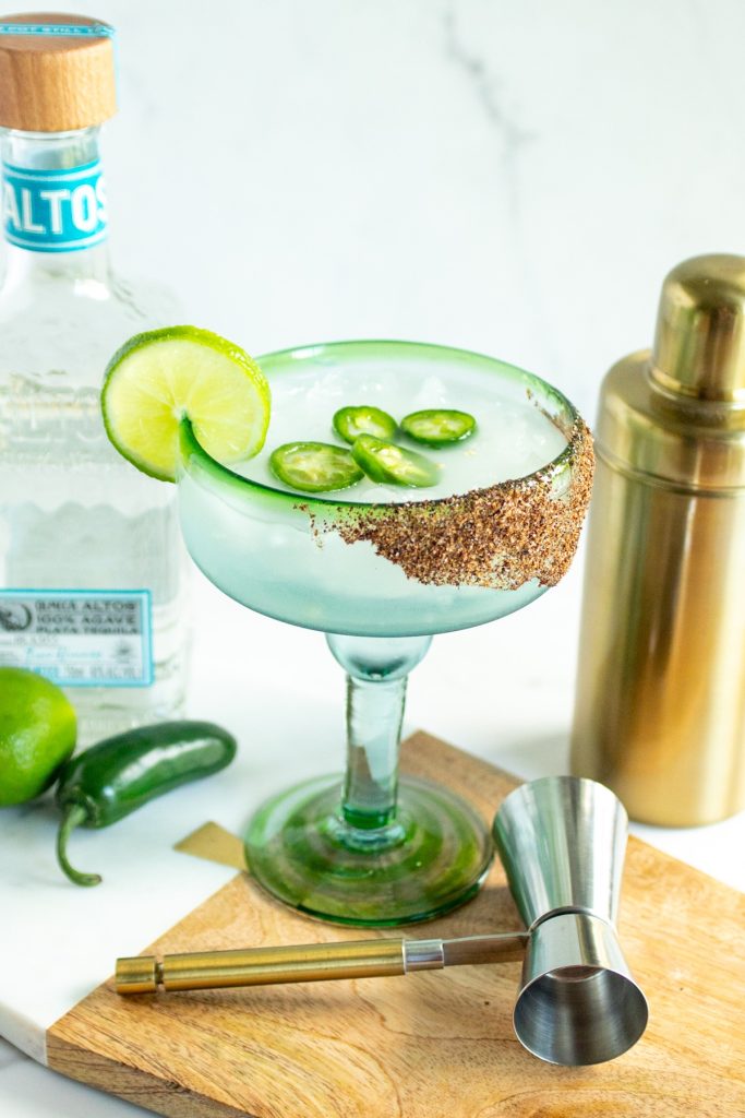 A margarita in a margarita-style glass garnished with jalapenos and lime. A bottle of tequila to the left. A gold cocktail shaker is to the right.