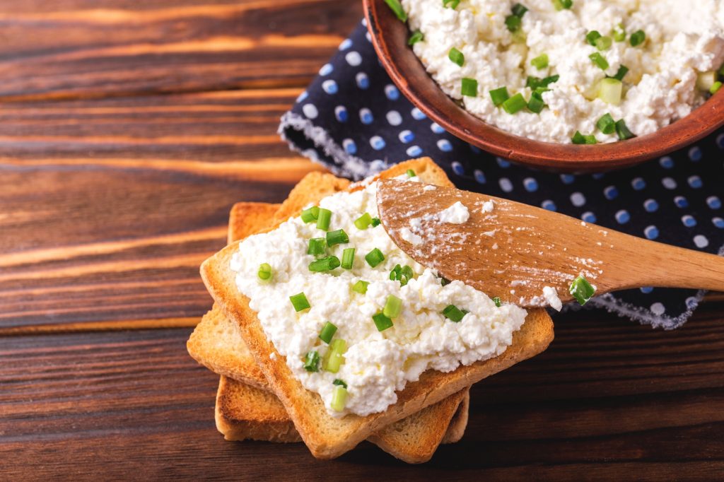 Cottage cheese on toast.  Wooden spreader.  Bowl of cottage cheese.