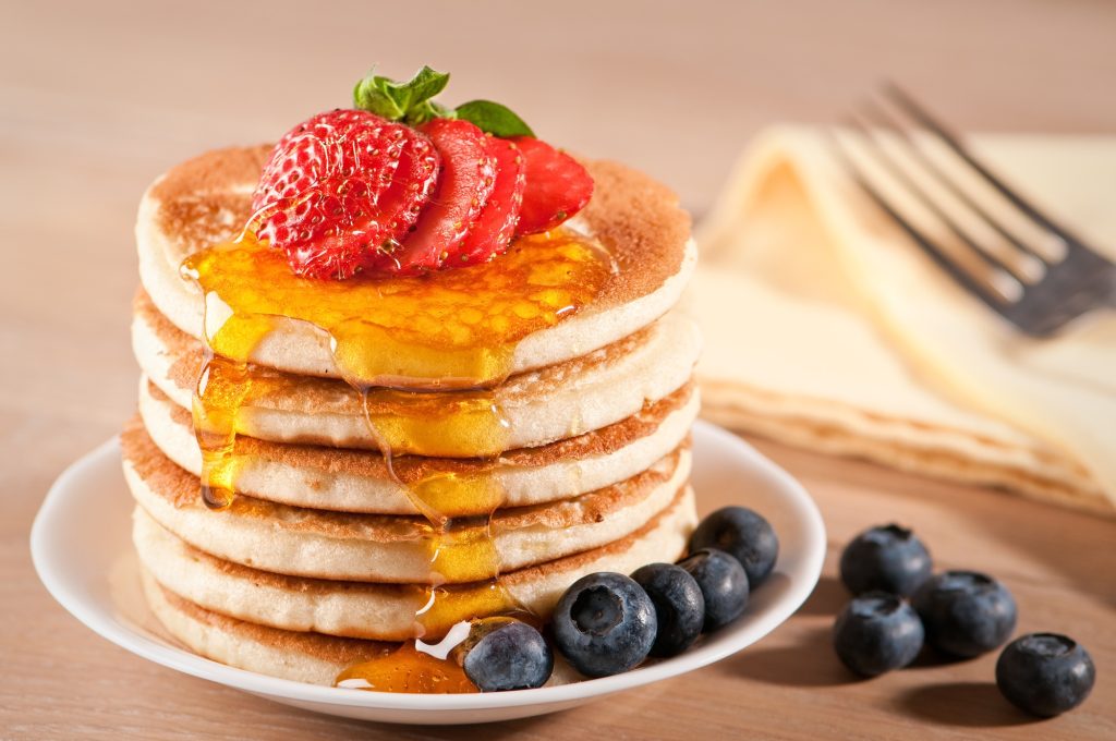 stack of pancakes on a plate with strawberries and blueberries. fork and cloth napkin to the right of plate.