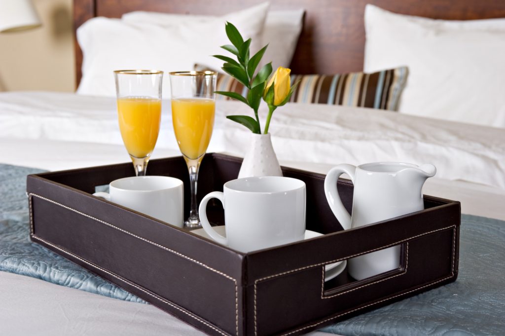 A on a hotel bed.  The tray has two flutes of orange juice and a vase of flowers.  Also has two white coffee cups.