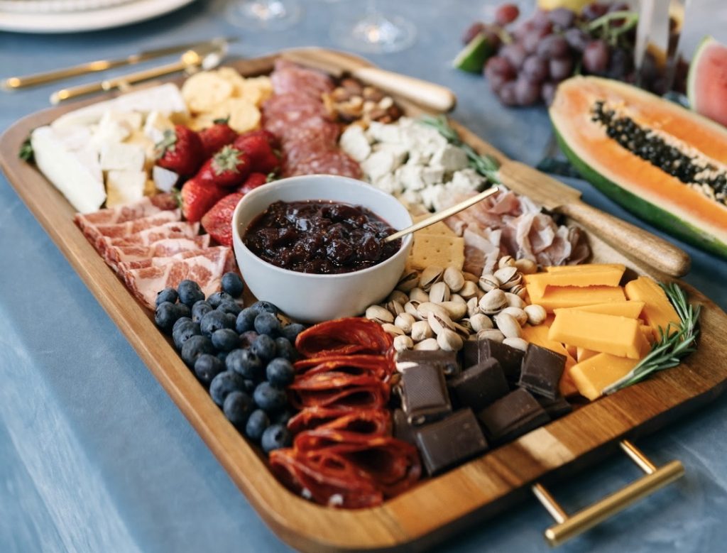 Charcuterie board with meats, cheeses, nuts, chocolate and crackers. It's on a wood serving tray with gold handles.