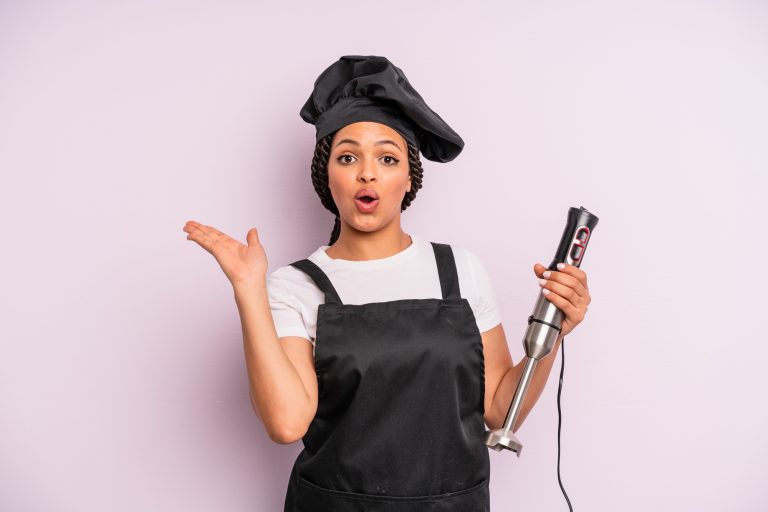 A woman smiling, holding an immersion blender.