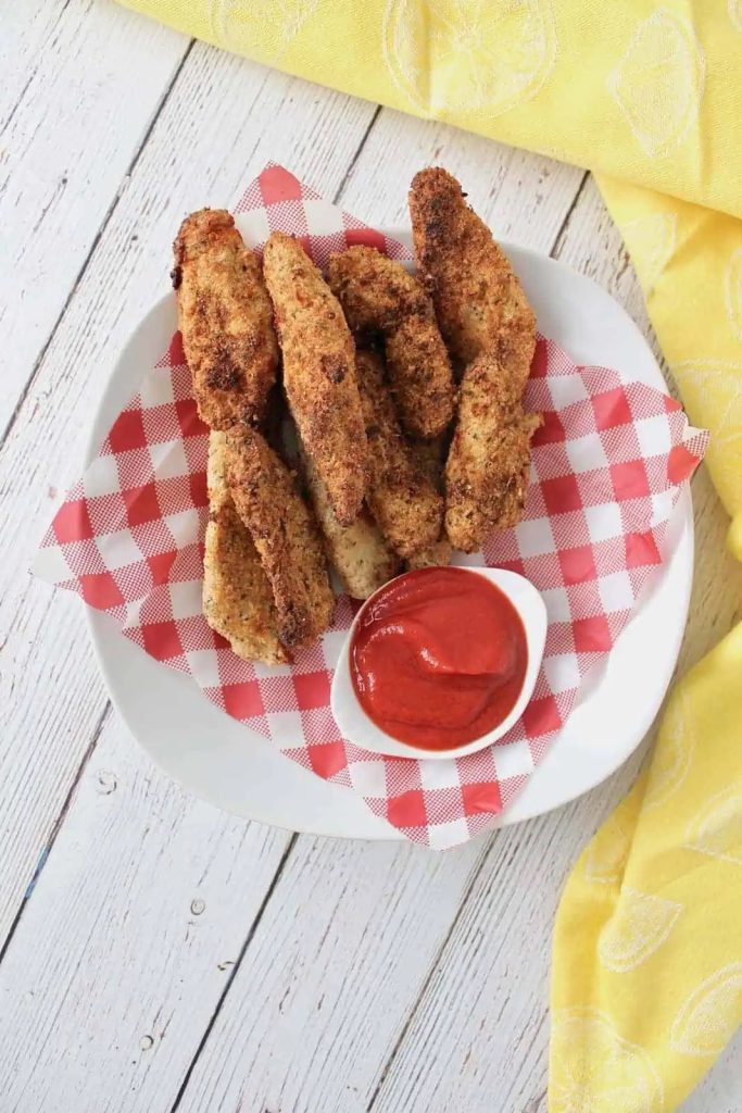 A whit plate of keto chicken tenders with a side of ketchup.  The backdrop is red and white gingham.