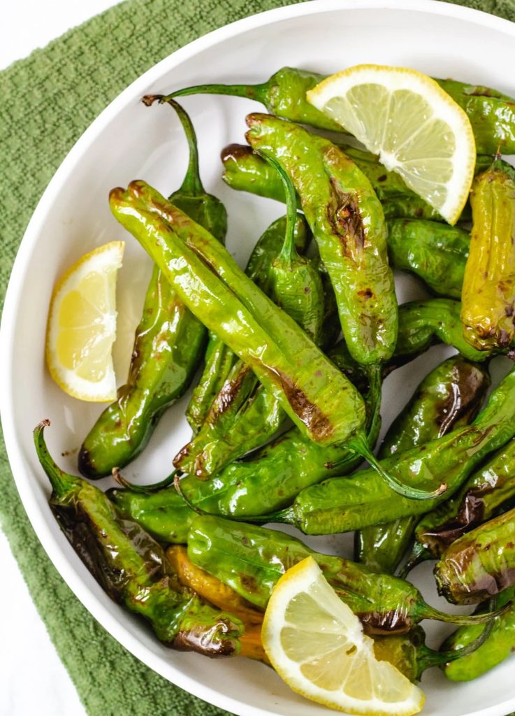 A white plate with air fried shishito peppers and wedges of lemon.