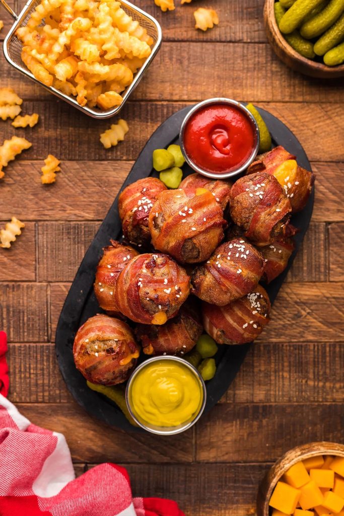 A black plate of bacon wrapped cheeseburger bites with cups of ketchup and mustard on the side.