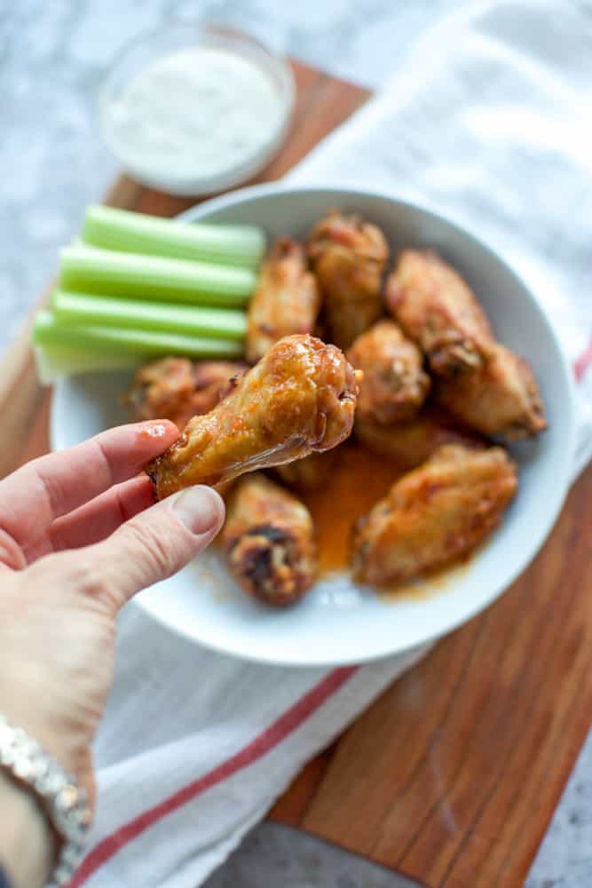 A bowl of buffalo wings with celery and ranch dressing. A hand holding one chicken wing.