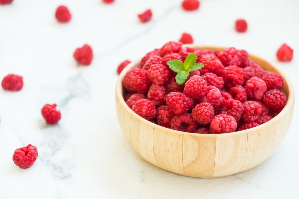 A light wooden bowl full of raspberries.  Serveral raspberries laying around the outside of the bowl.