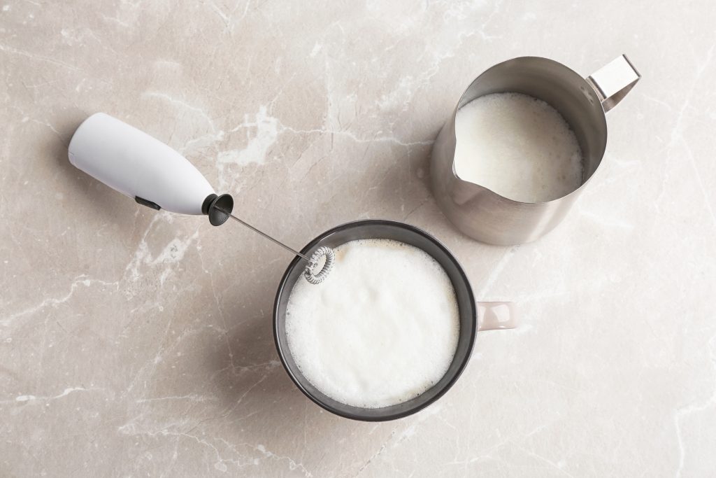 A battery operated frother and a cup of coffee, and a cup of frothed milk.